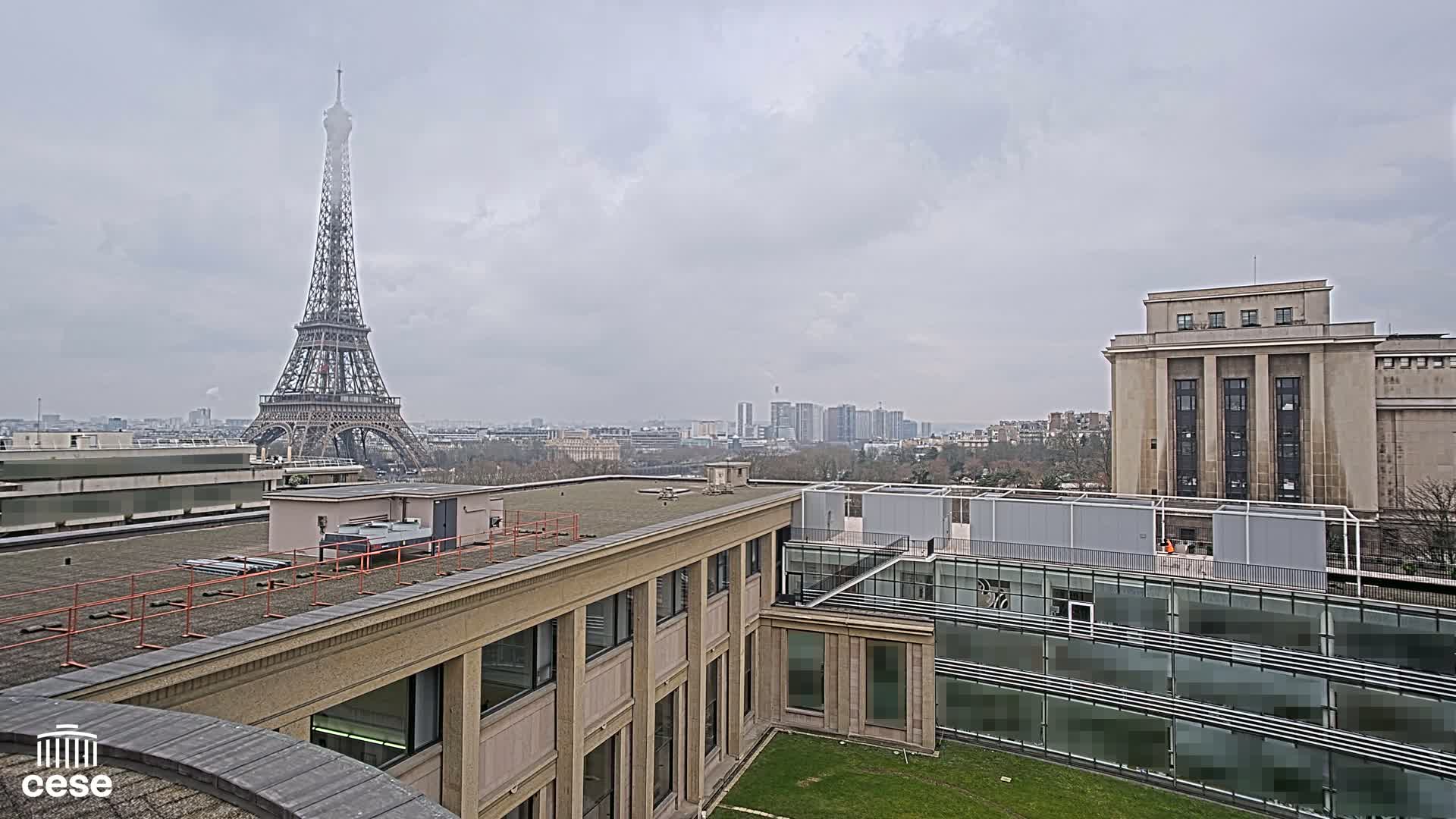 A rooftop view on an overcast day shows the Eiffel Tower in the distance, alongside other buildings, including a large tan structure with a flat roof and modern glass additions.