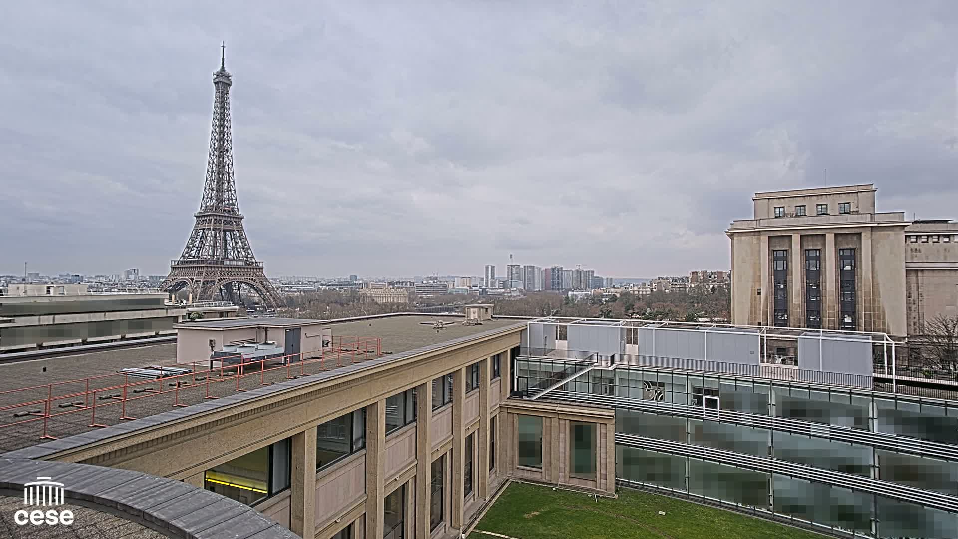 A view from a rooftop shows the Eiffel Tower in the distance under a cloudy sky, with a modern building in the foreground and a large classical-style building to the right.