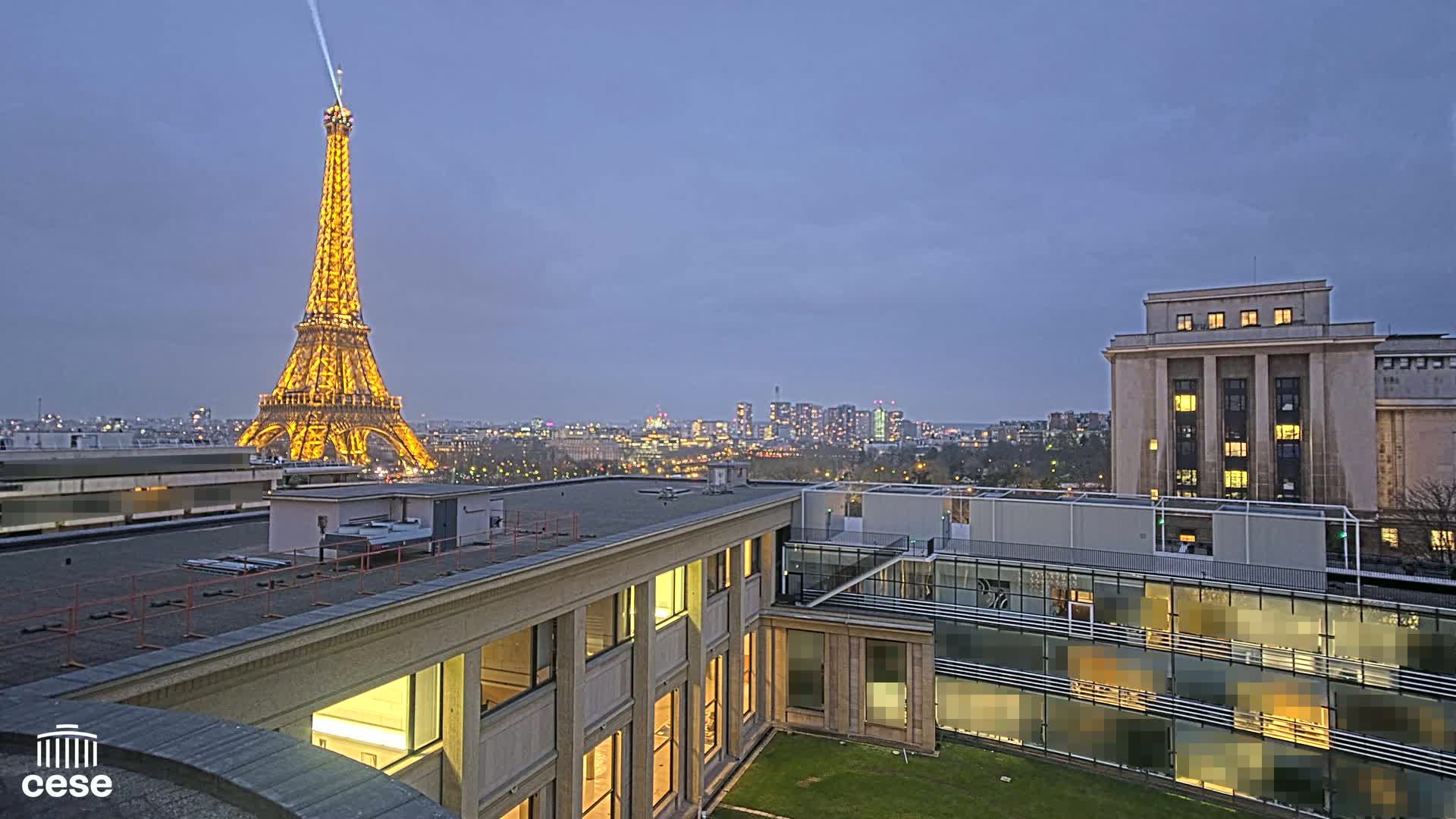 A brightly lit Eiffel Tower is visible in the distance from a rooftop vantage point overlooking a cityscape at dusk.