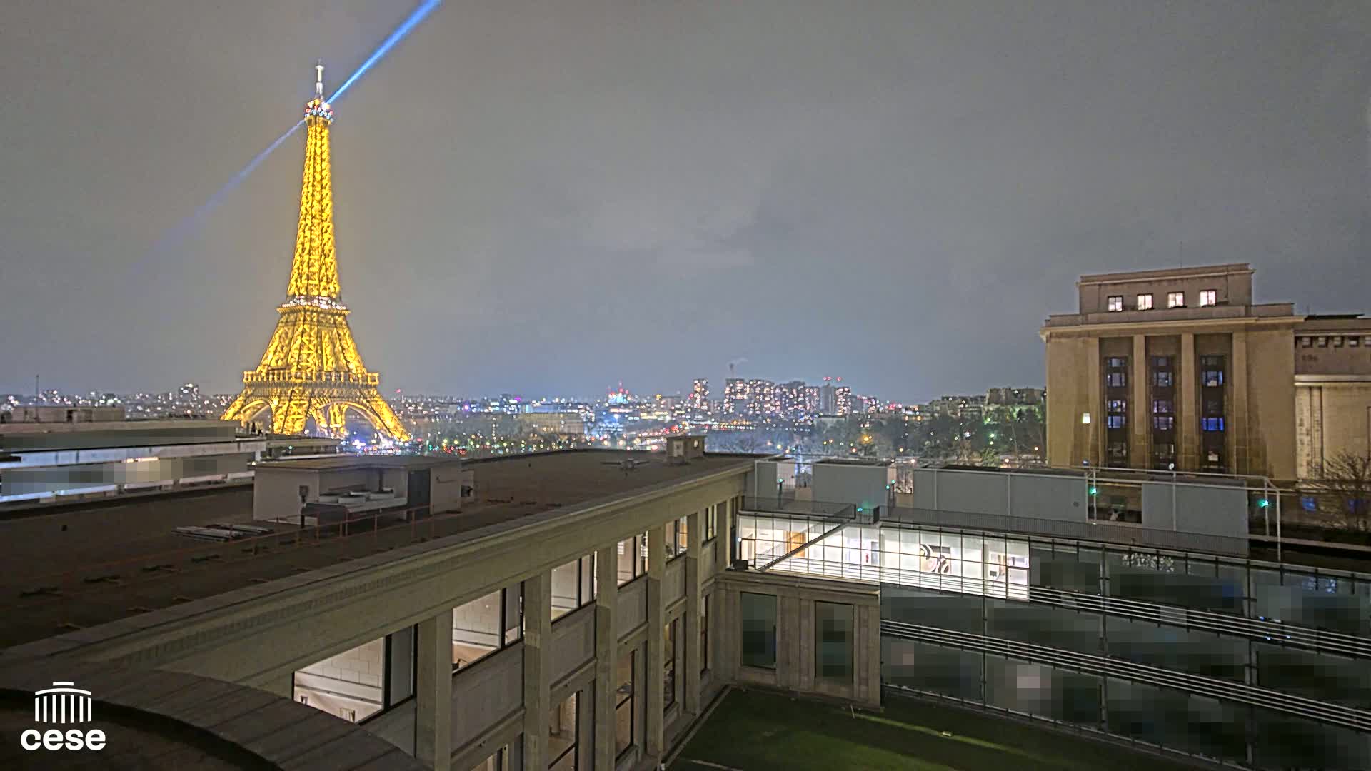 A nighttime view of the Eiffel Tower illuminated, a city skyline, and several buildings, under a cloudy sky.