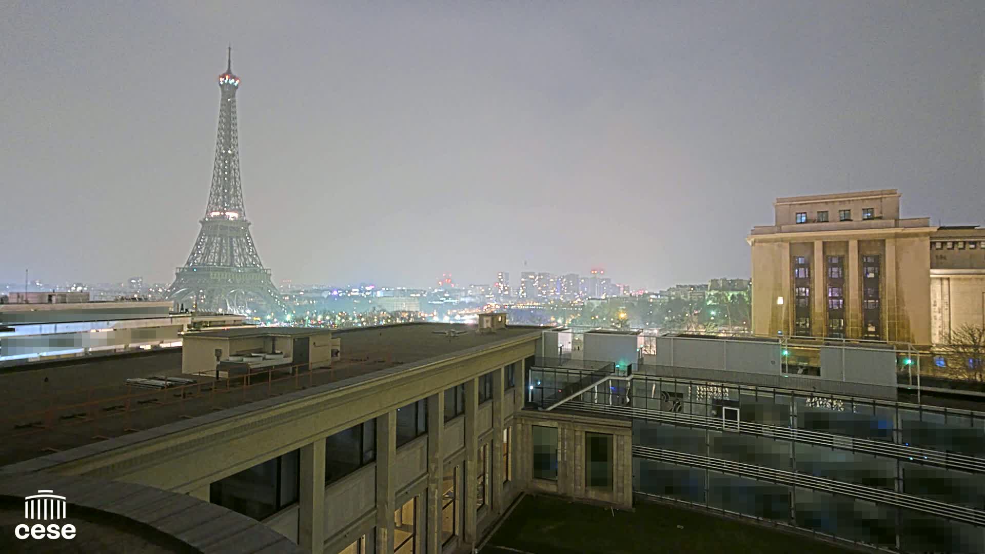 A nighttime view of the Eiffel Tower in a hazy atmosphere, seen from a rooftop overlooking a city with other buildings.