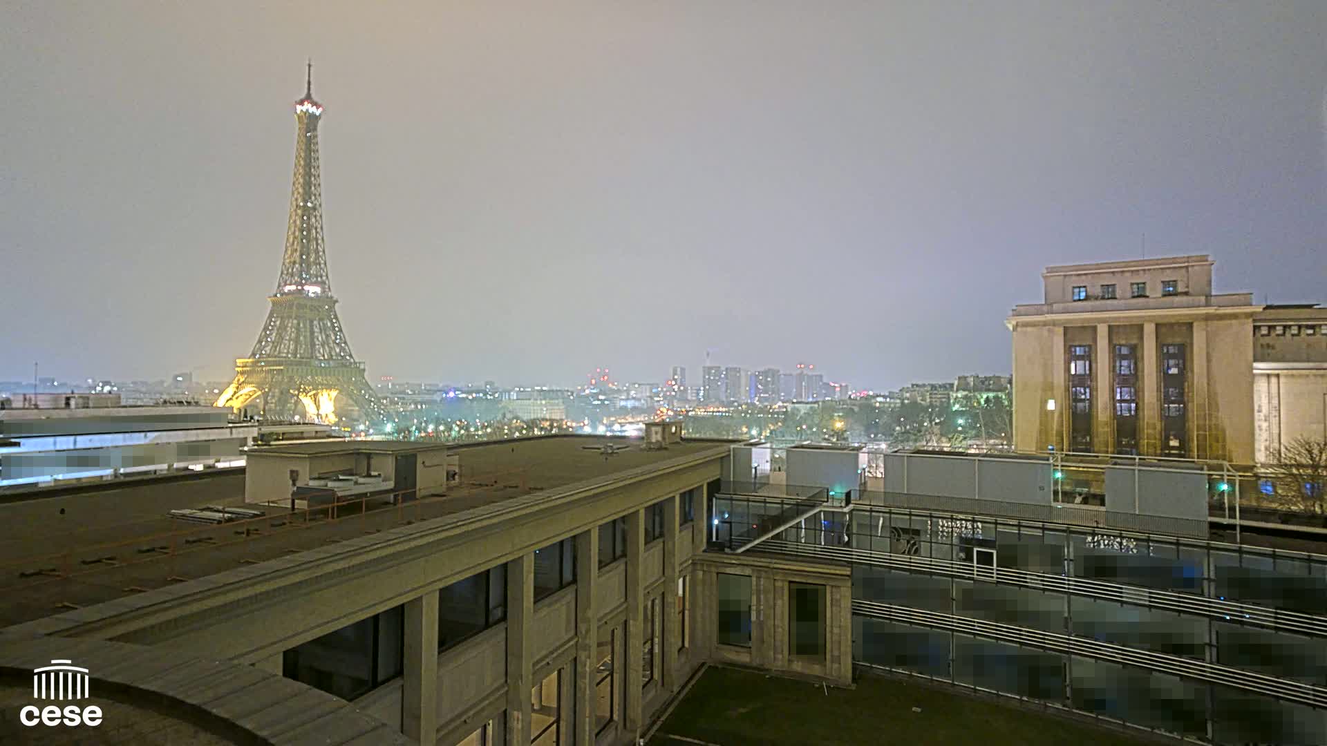 A nighttime view of the Eiffel Tower in a hazy atmosphere, with city lights visible in the distance and a large building in the foreground.