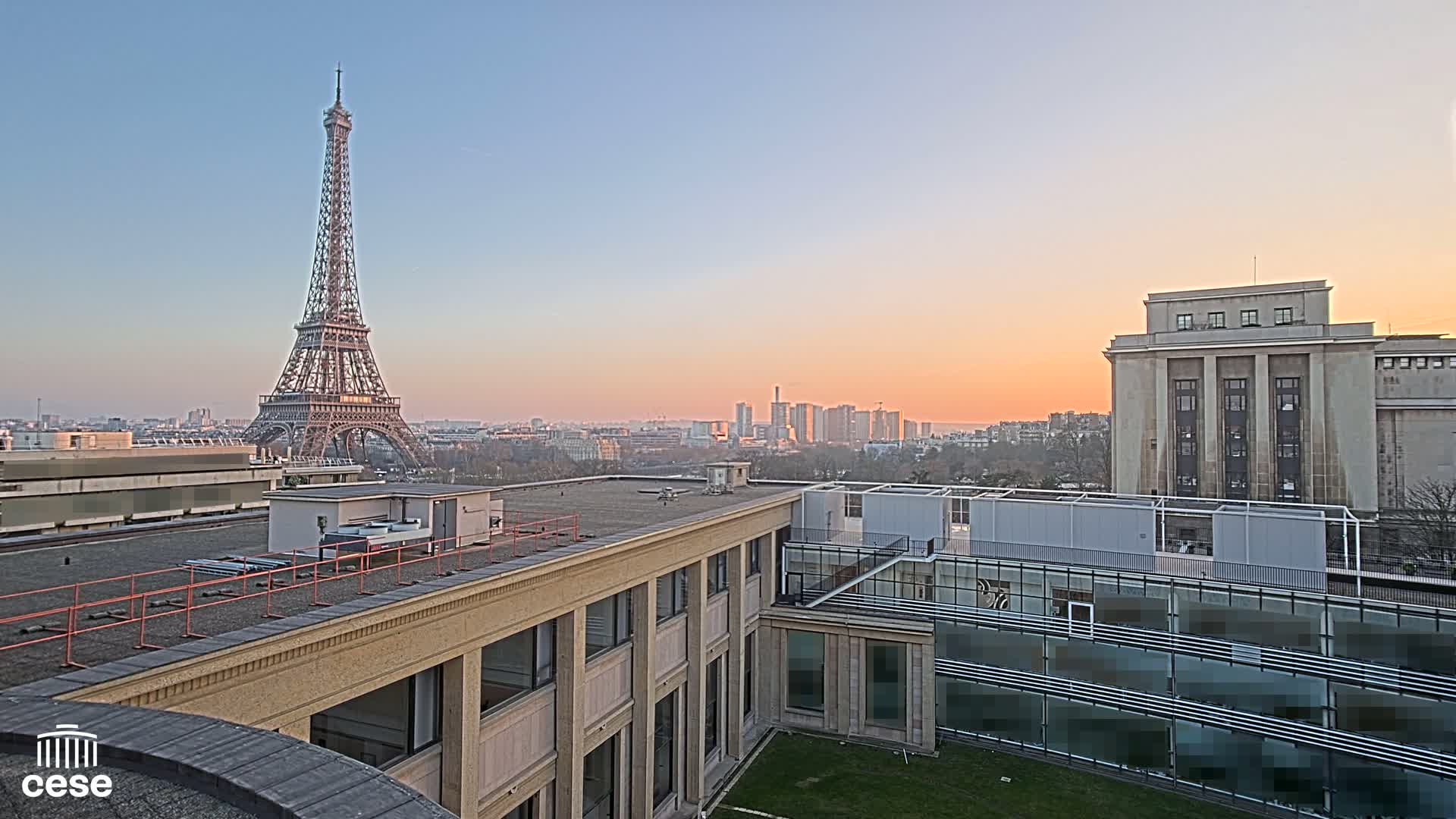 A rooftop view of the Eiffel Tower at sunset, overlooking a cityscape and a large, light-colored building.
