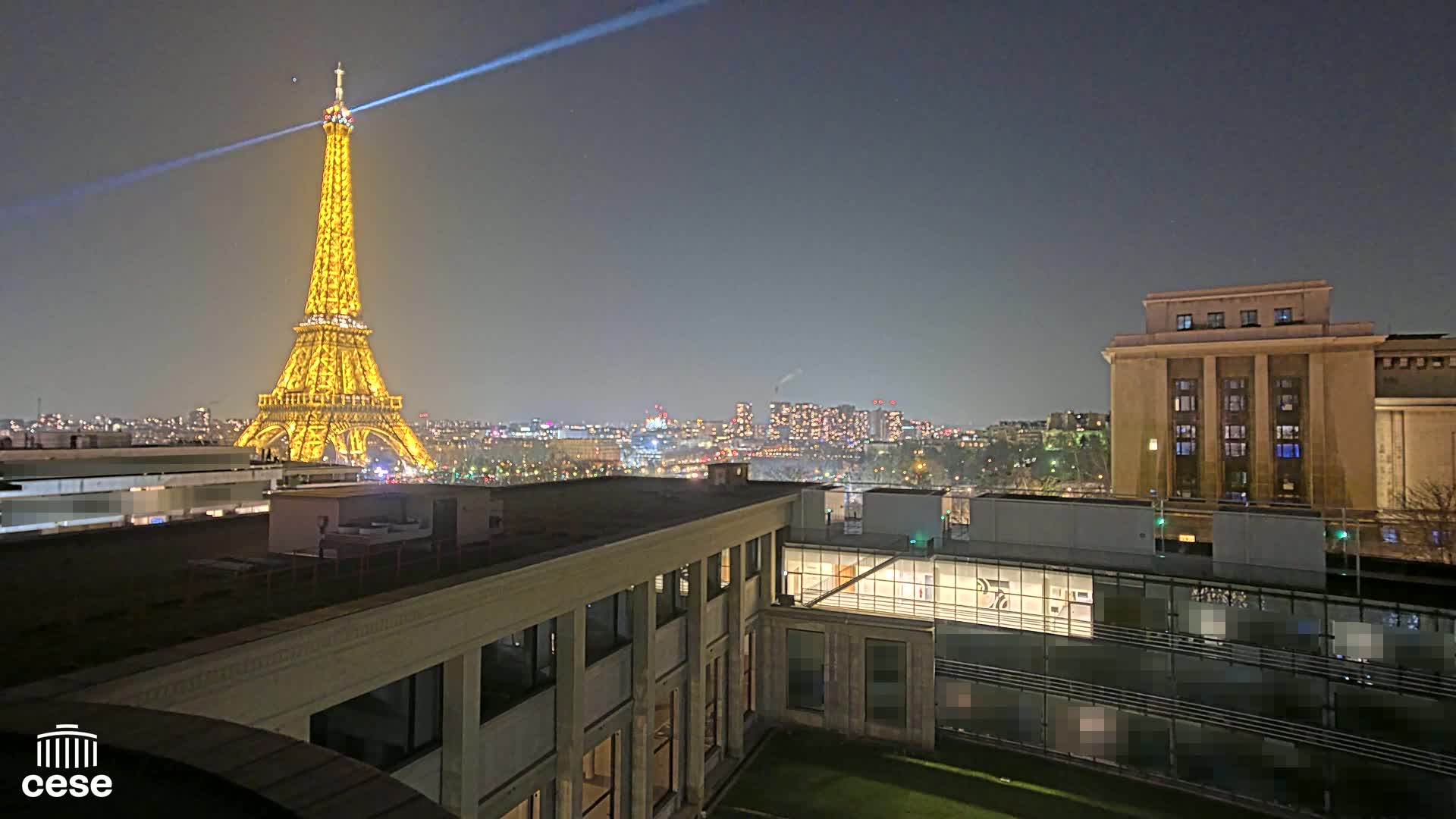 A nighttime view of the Eiffel Tower, illuminated in gold, with a city skyline in the background and buildings in the foreground under a clear, dark sky.