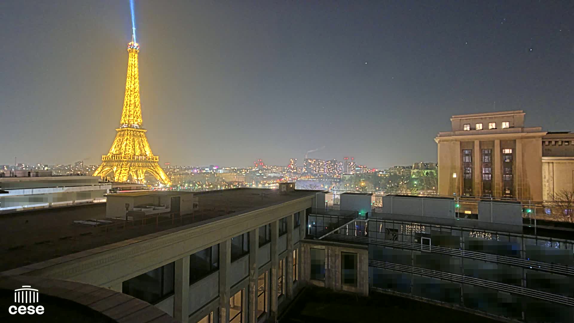 A nighttime view from a rooftop shows the Eiffel Tower illuminated, a city skyline, and a large building in the foreground under a clear, dark sky.
