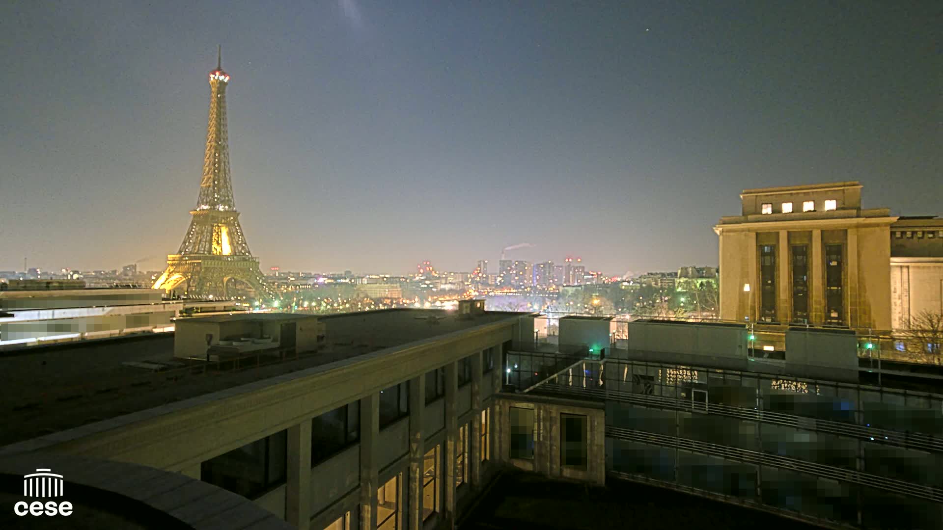 A nighttime view from a rooftop shows the Eiffel Tower illuminated in the distance, with a city skyline and a large building in the foreground under a clear, dark sky.