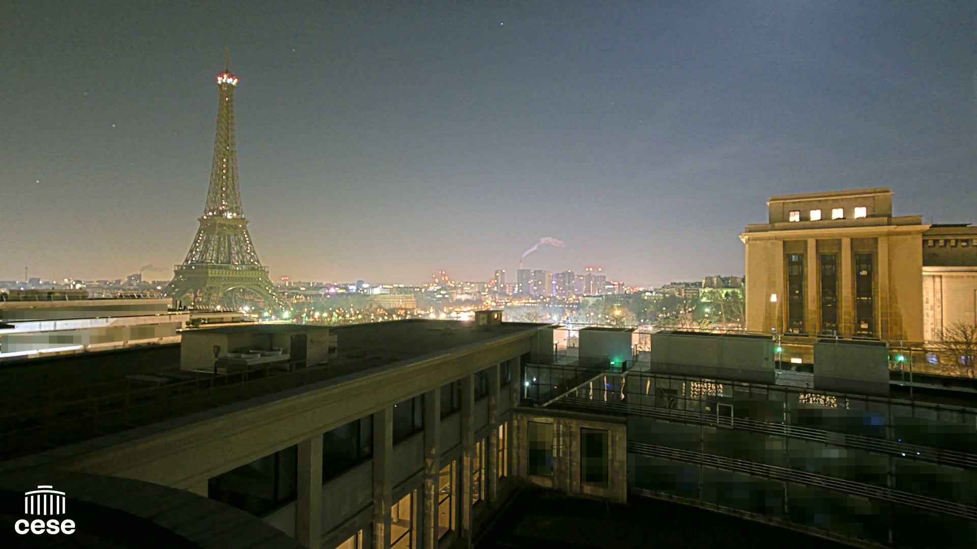 A nighttime view from a rooftop shows the Eiffel Tower and a city skyline under a mostly clear sky.