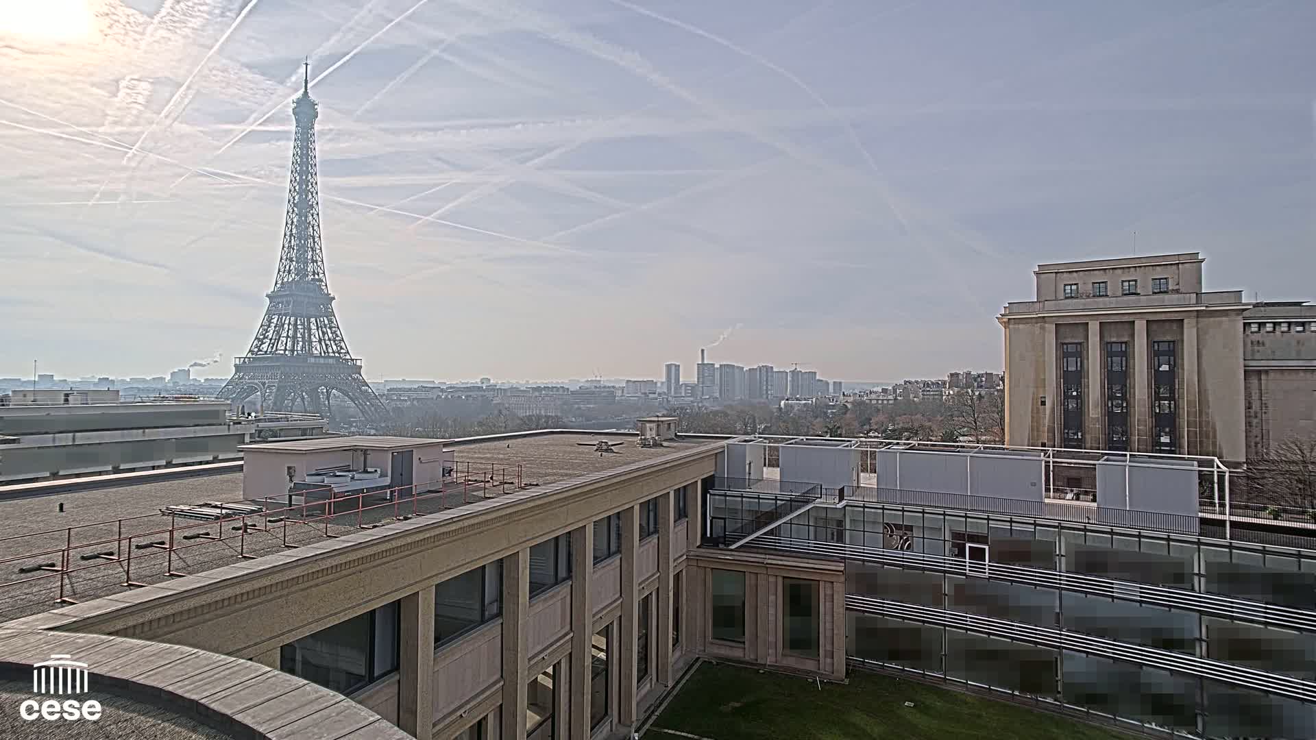 A distant view of the Eiffel Tower on a hazy day, seen from atop a building complex with a flat roof, adjacent to another large building.