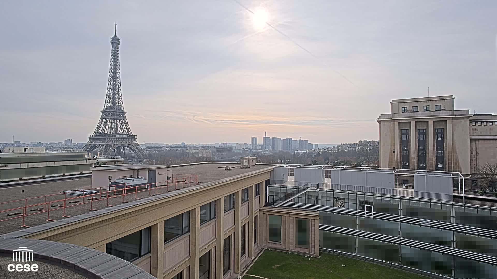 A rooftop view shows the Eiffel Tower in the distance on a partly sunny day, with city buildings and a grassy courtyard in the foreground.