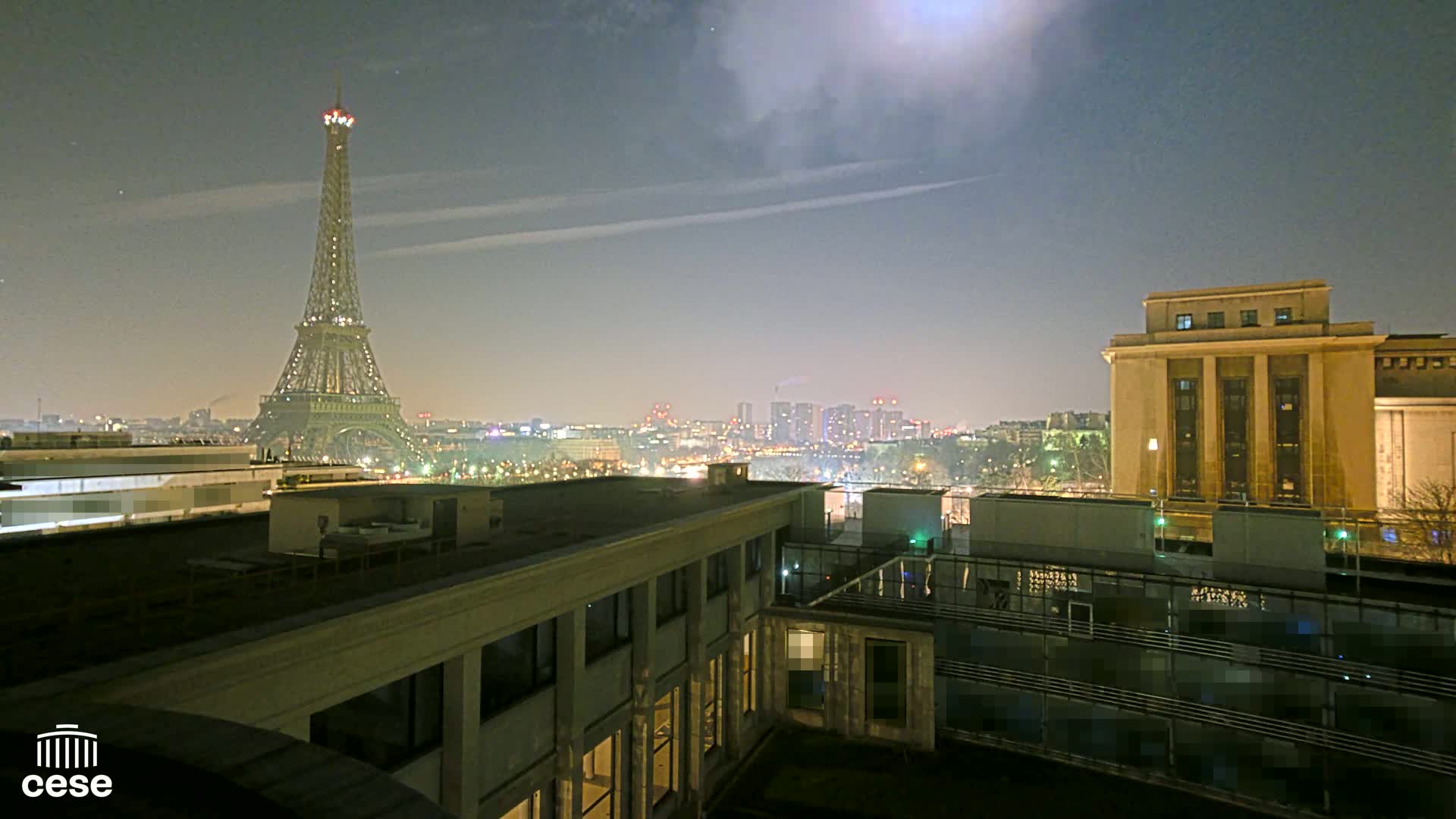 A nighttime view of the Eiffel Tower from a rooftop, with a hazy, moonlit sky and city lights in the distance.