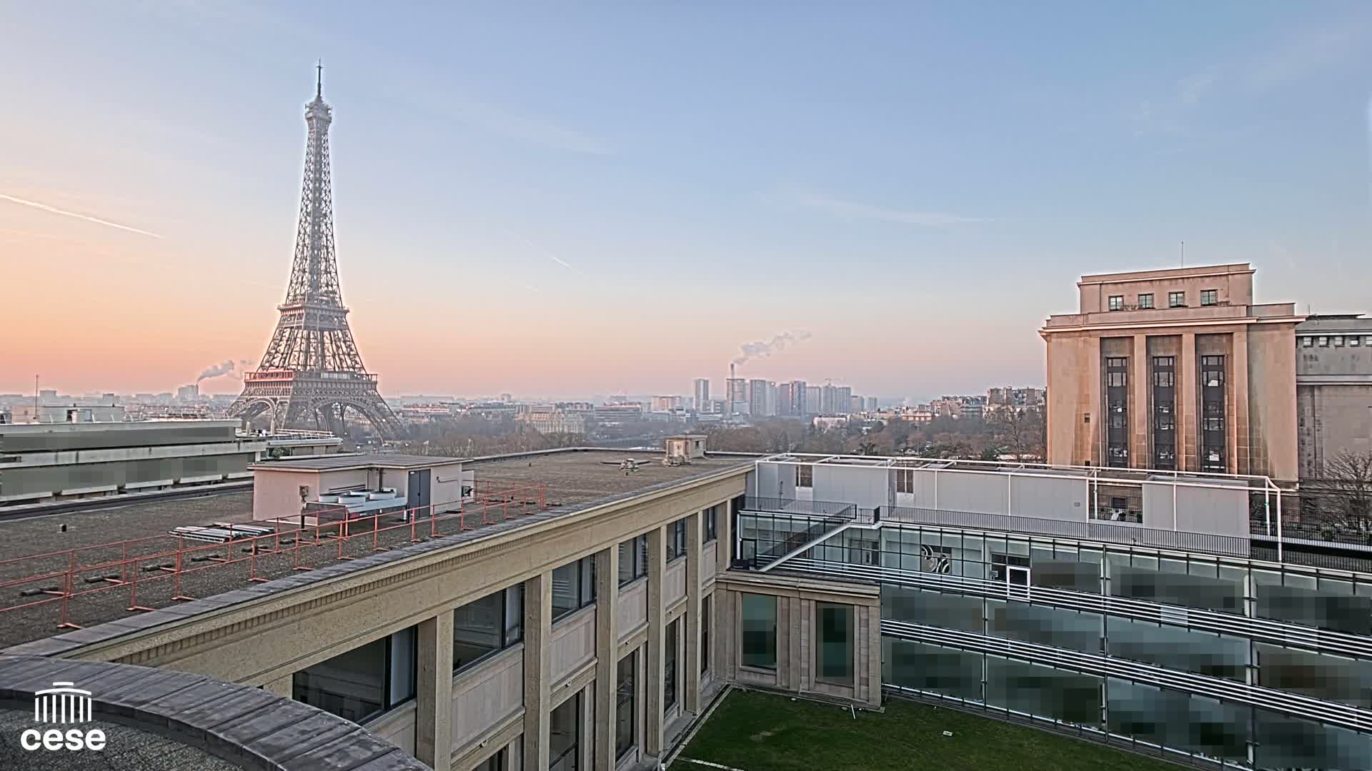 A Parisian rooftop view at dawn shows the Eiffel Tower in the distance, with city buildings and a pale pink sky.