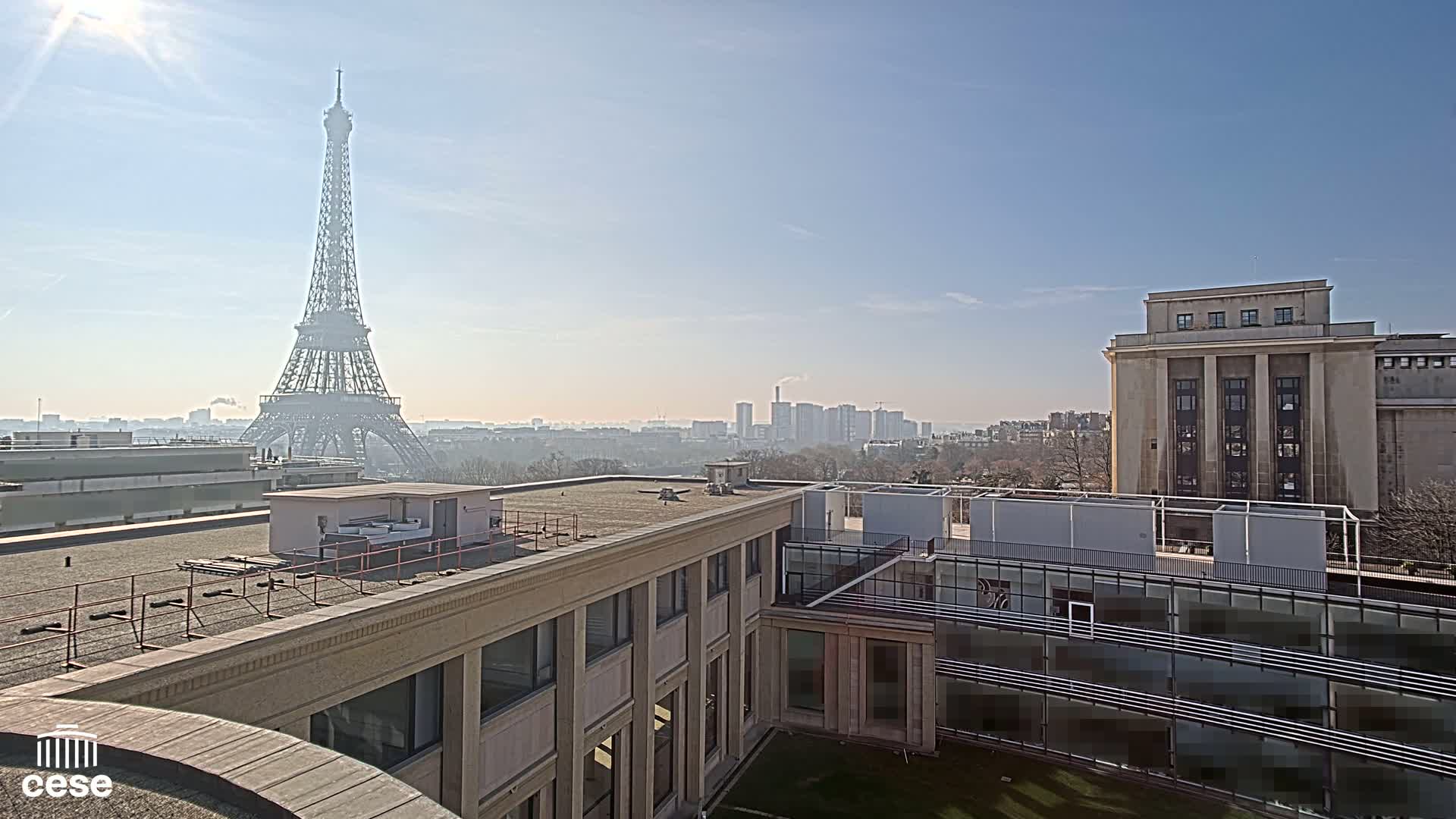 A sunny view from a rooftop shows the Eiffel Tower in the distance, city buildings, and a courtyard with modern architectural features.