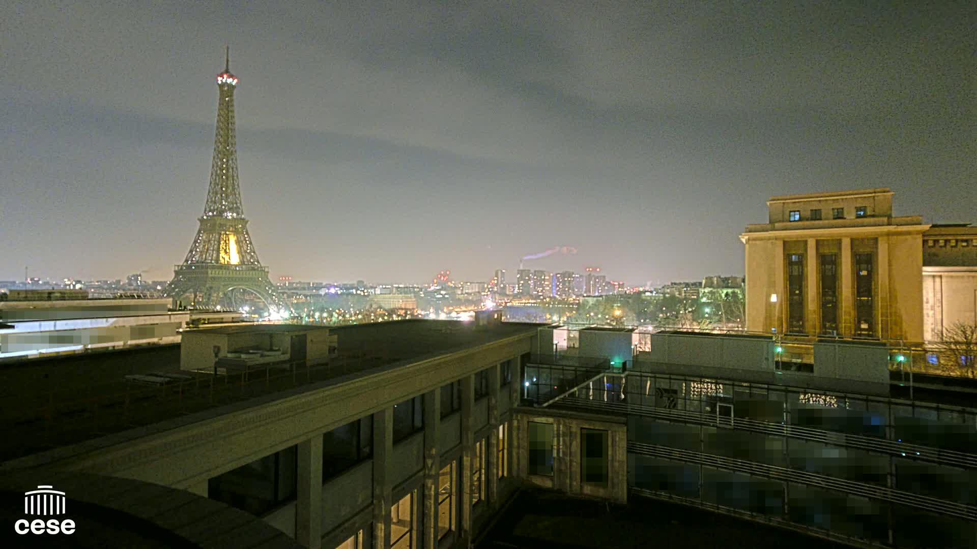 A nighttime cityscape with the Eiffel Tower in the distance, viewed from a rooftop under a hazy sky.