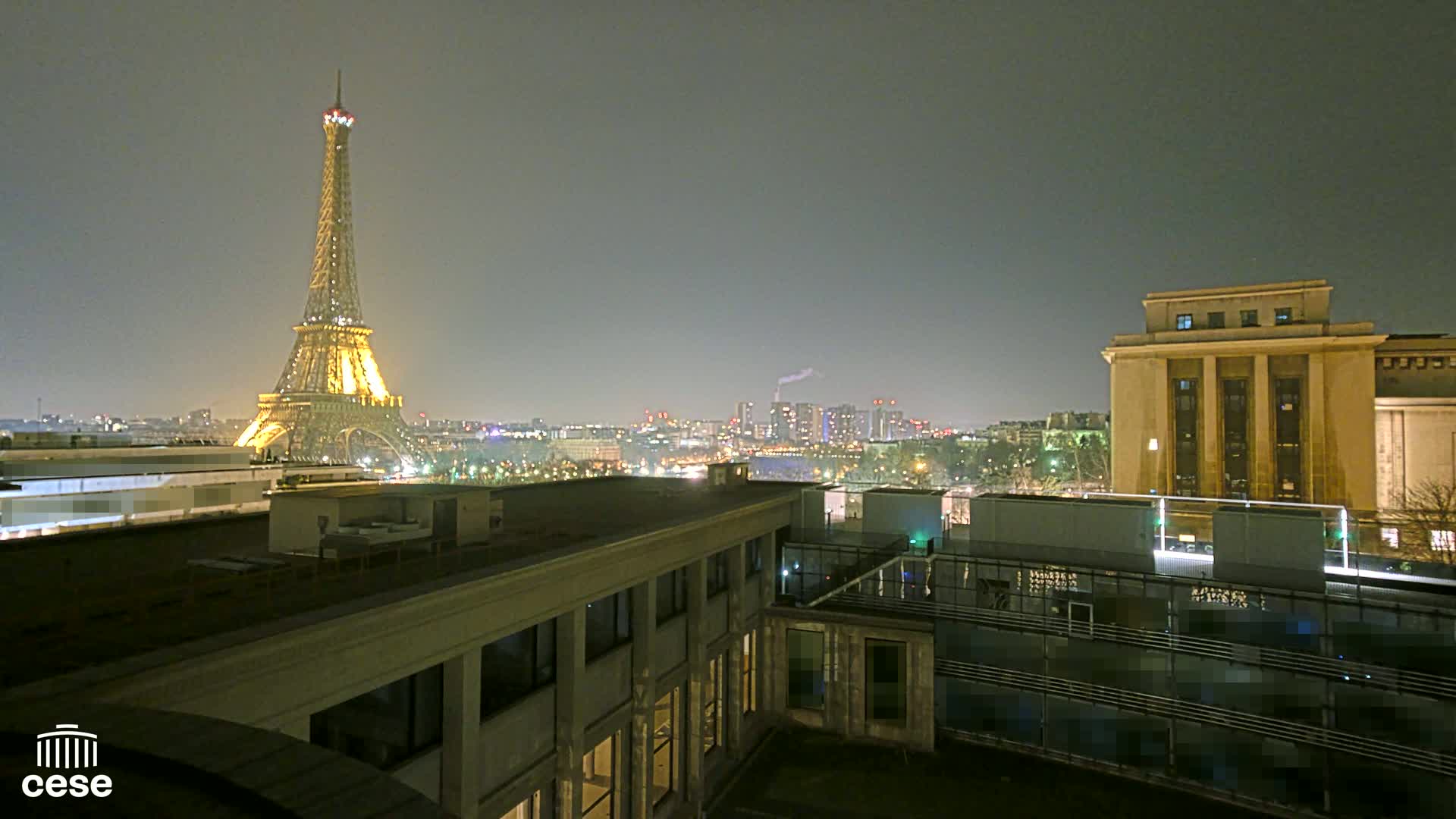 A nighttime cityscape view shows the Eiffel Tower illuminated, a city skyline in the distance, and a portion of a building's rooftop in the foreground.
