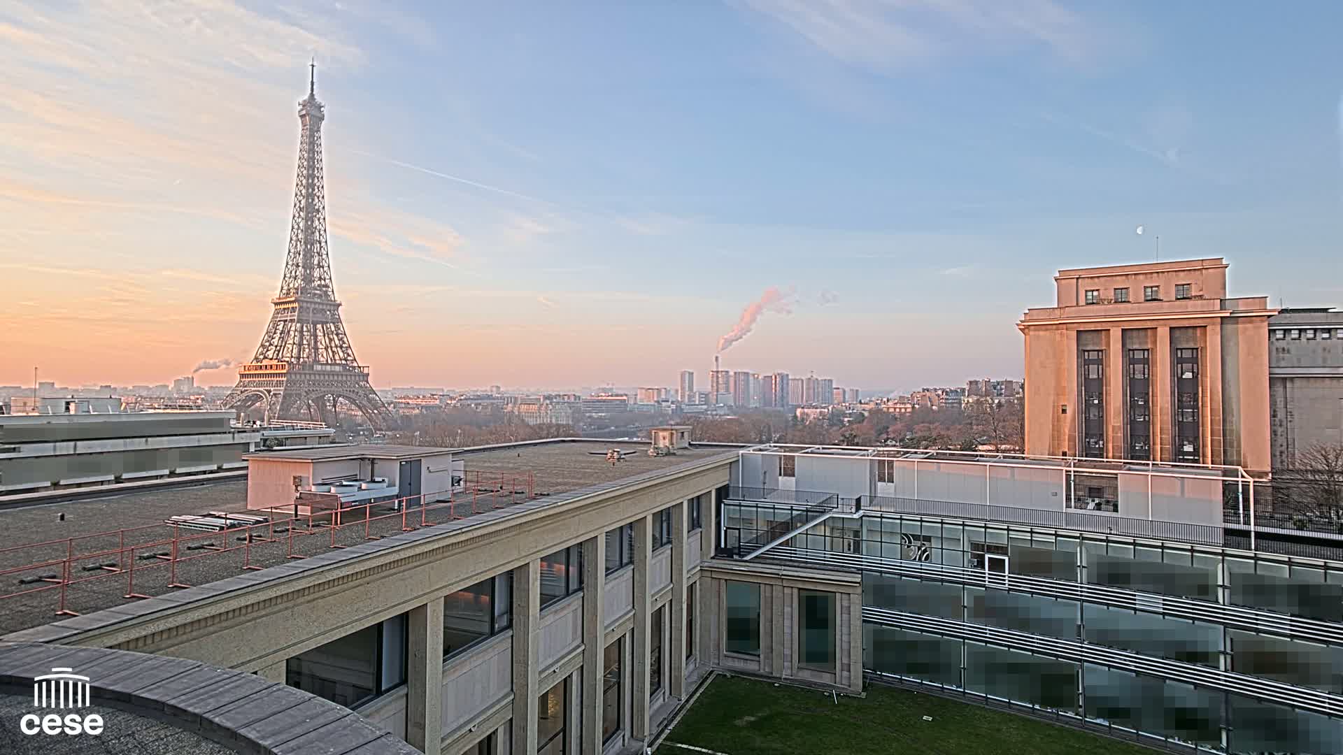 A cityscape at sunrise features the Eiffel Tower in the distance, a large building in the foreground, and a hazy sky with a plume of smoke rising from the city.