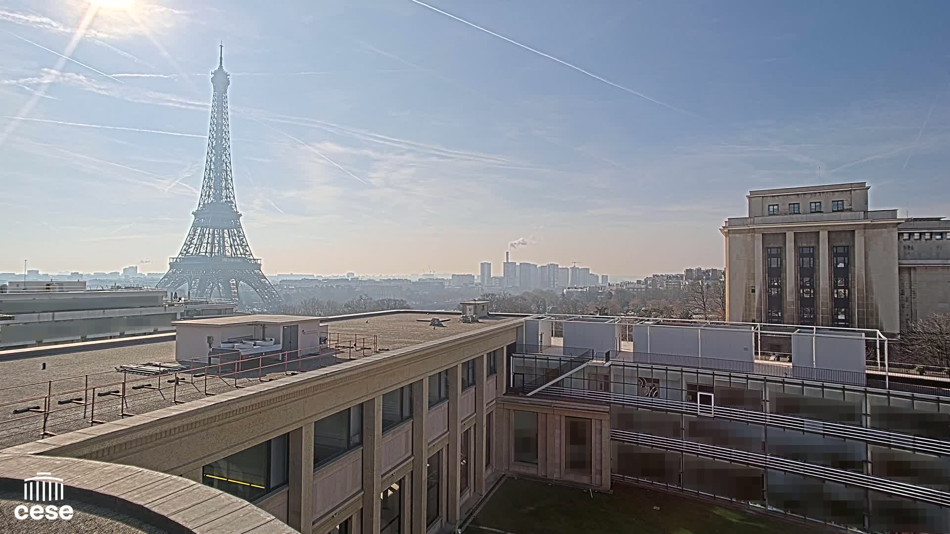 A distant view of the Eiffel Tower on a sunny day, seen from a rooftop overlooking other buildings and a cityscape.