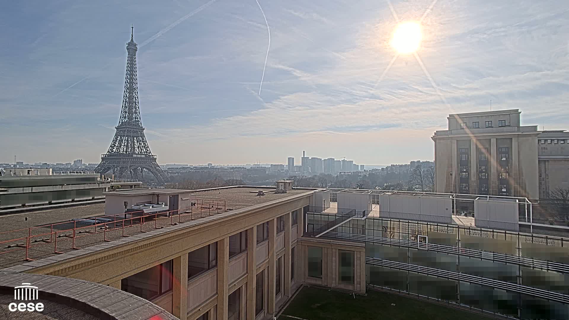 A sunny day reveals the Eiffel Tower in the distance, viewed from a rooftop overlooking a city skyline and a large, light-colored building.