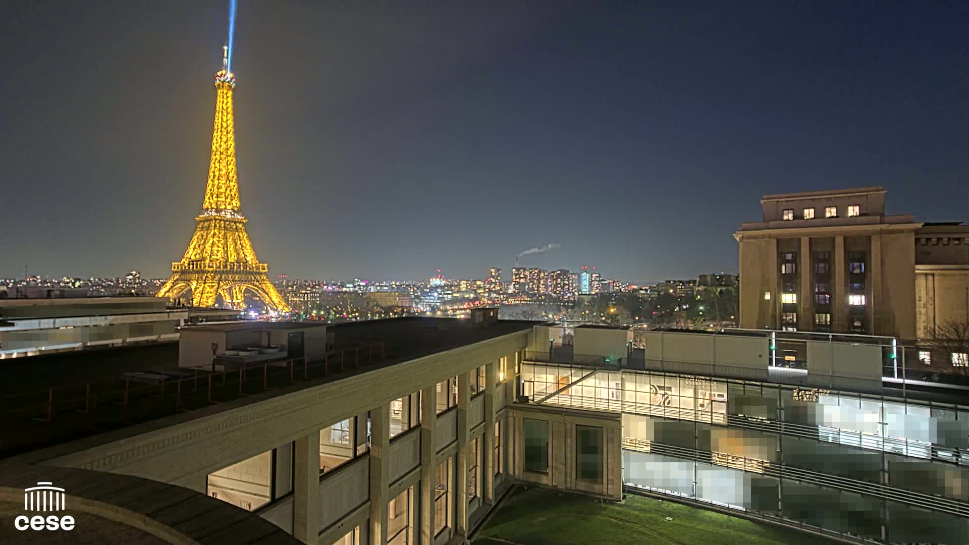 A nighttime view of the Eiffel Tower illuminated in gold, a city skyline, and a modern building complex under a clear, dark sky.