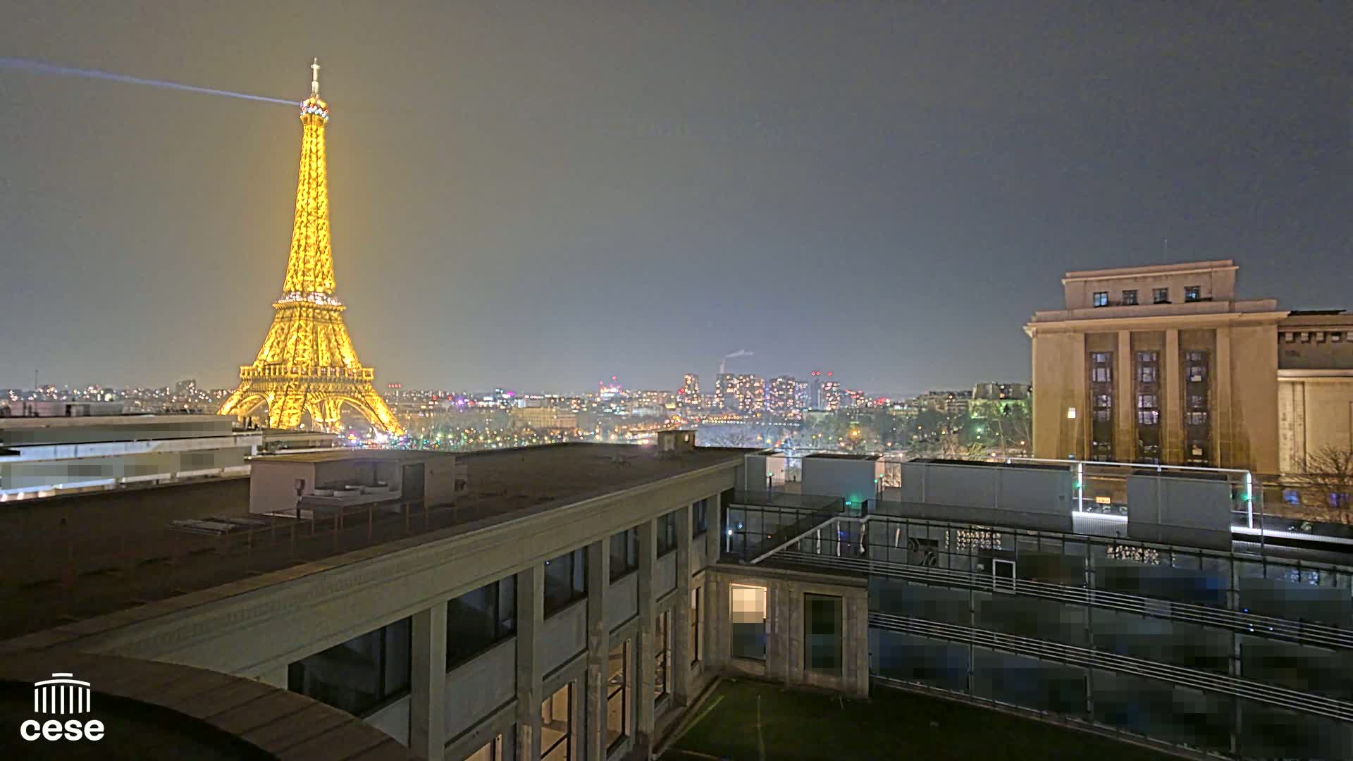 A nighttime view of the Eiffel Tower illuminated, along with a cityscape and nearby buildings under a hazy sky.