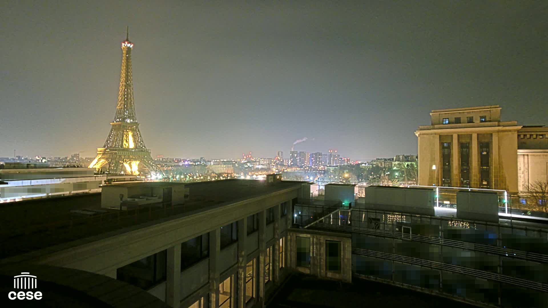 A nighttime view of the Eiffel Tower illuminated, a city skyline, and a large building, under a hazy sky.