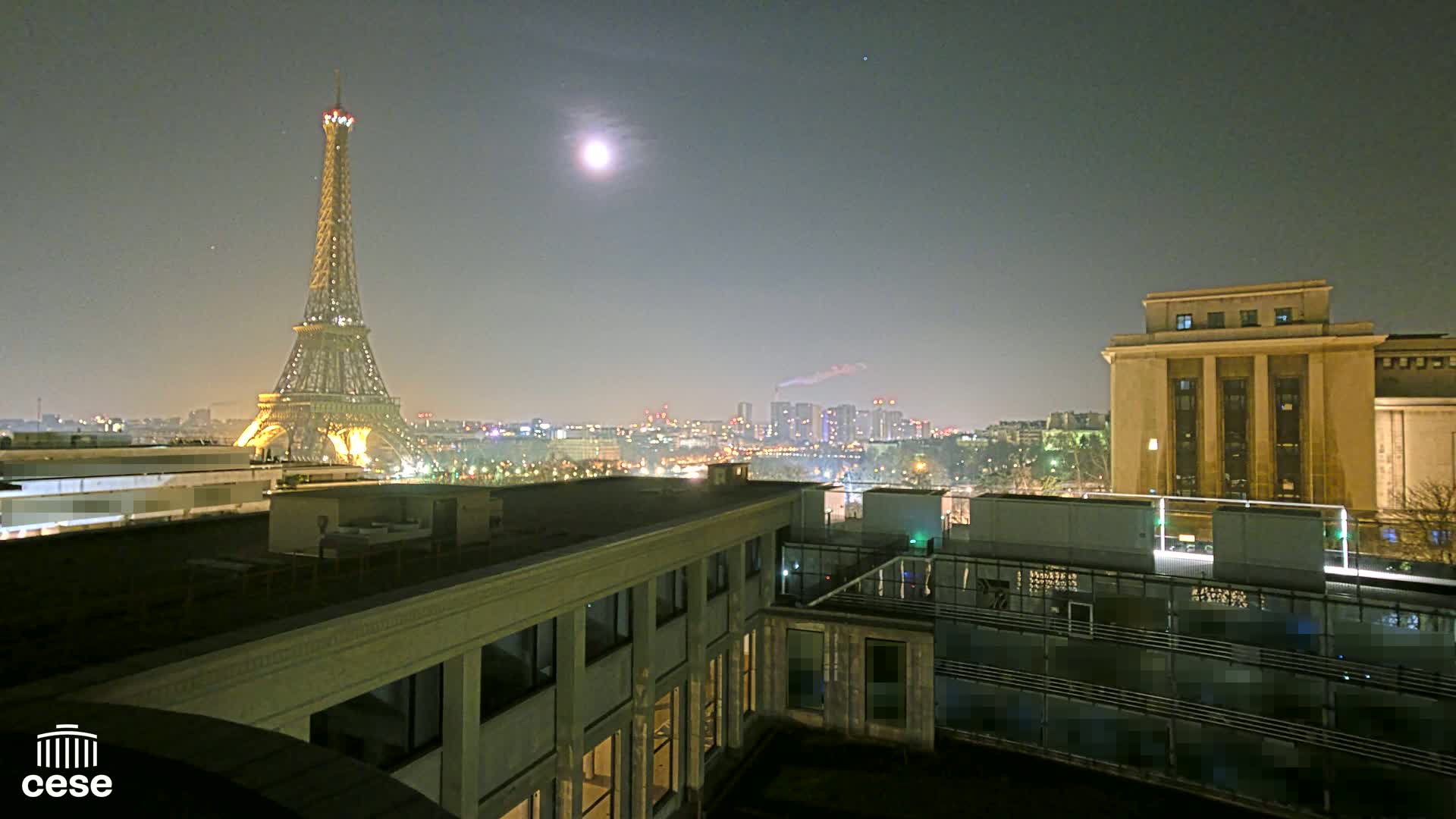 A nighttime view of the Eiffel Tower, illuminated against a dark sky with a bright moon, overlooking a city skyline and a large building.
