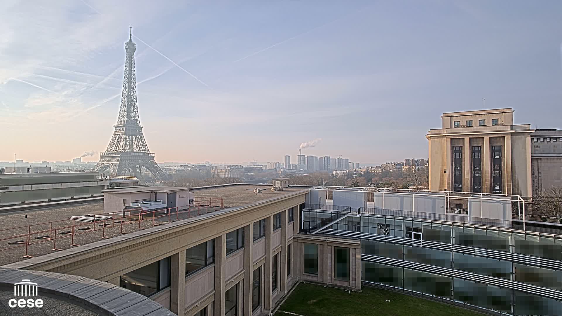 A cityscape view shows the Eiffel Tower in the distance under a hazy, bright sky, with several buildings in the foreground.