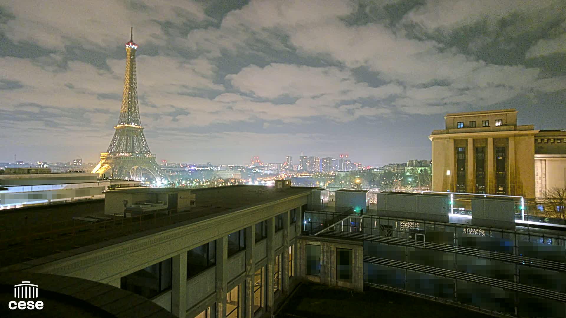 A nighttime cityscape view shows the Eiffel Tower illuminated against a cloudy sky, with a large building and city lights in the foreground.
