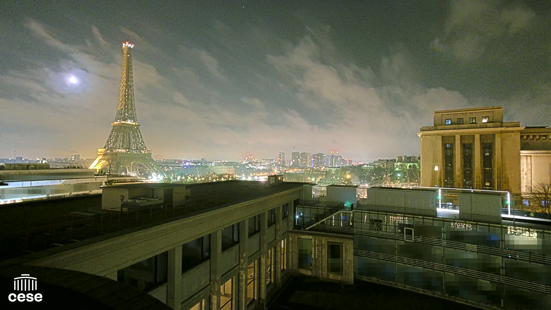 A nighttime view of the Eiffel Tower illuminated against a cloudy sky, with city lights and a large building in the foreground.
