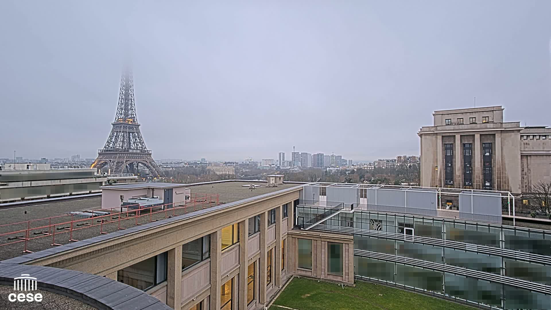 A foggy cityscape is seen from a rooftop, featuring the Eiffel Tower in the distance and modern buildings in the foreground.