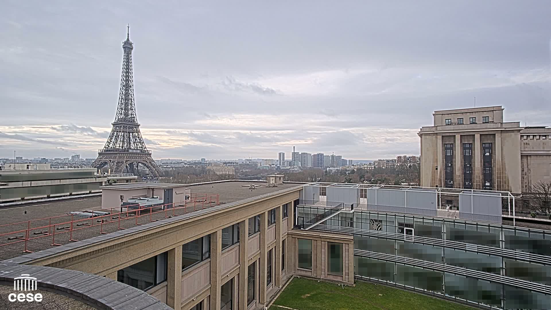 A rooftop view of Paris on an overcast day shows the Eiffel Tower in the distance, with modern and classical buildings in the foreground.