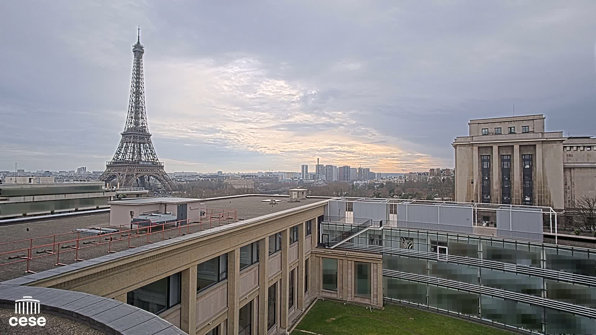 A view from a rooftop shows the Eiffel Tower in the distance under a cloudy sky, with other buildings and a cityscape visible in the background.