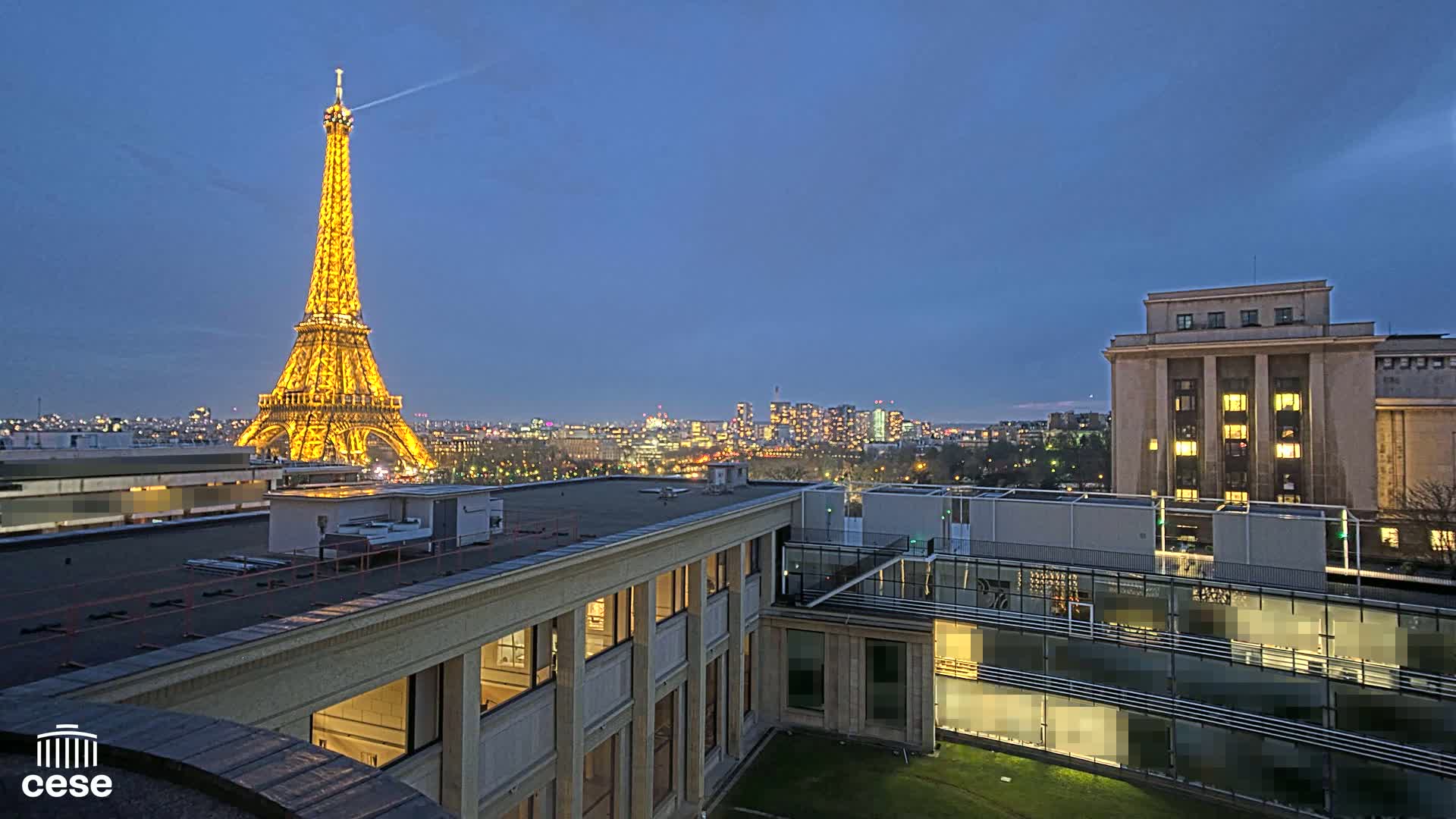 The illuminated Eiffel Tower is visible at dusk from a rooftop overlooking a city skyline and a modern building.