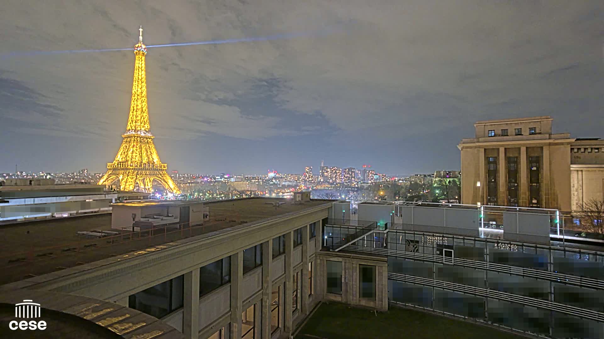 A nighttime view of the Eiffel Tower illuminated against a cloudy sky, seen from a rooftop overlooking a city skyline and another building.