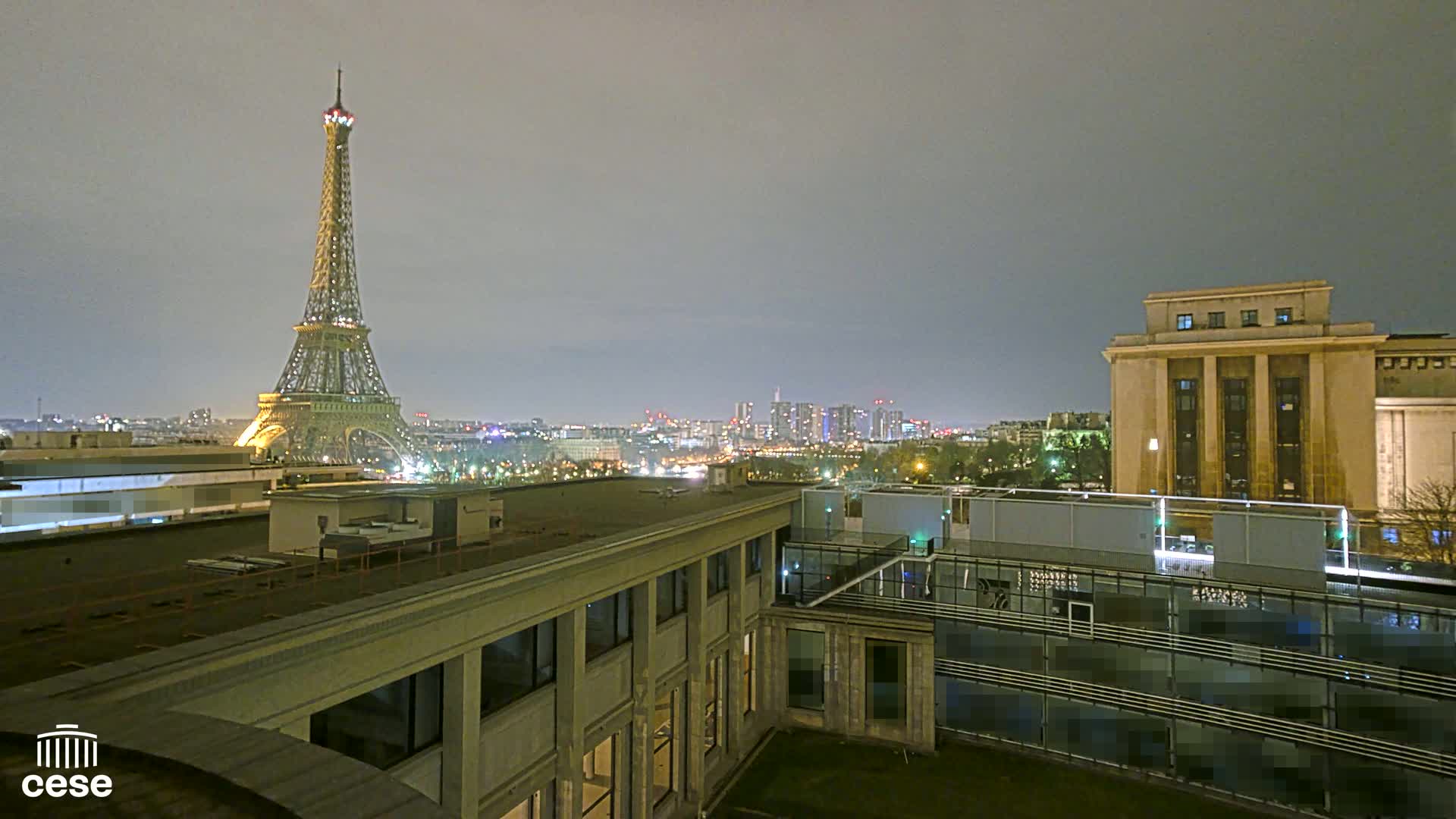 A nighttime cityscape view of the Eiffel Tower and Paris skyline from a rooftop, under a cloudy sky.