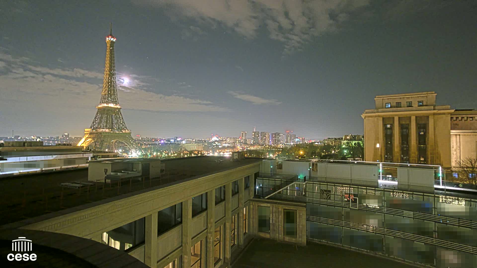 A nighttime view of the Eiffel Tower illuminated against a partly cloudy sky, from a rooftop overlooking a city skyline and a large building.