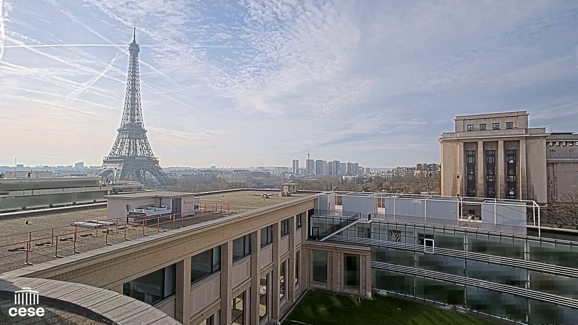 A rooftop view of Paris on a partly cloudy day shows the Eiffel Tower in the distance, along with other buildings and a cityscape.