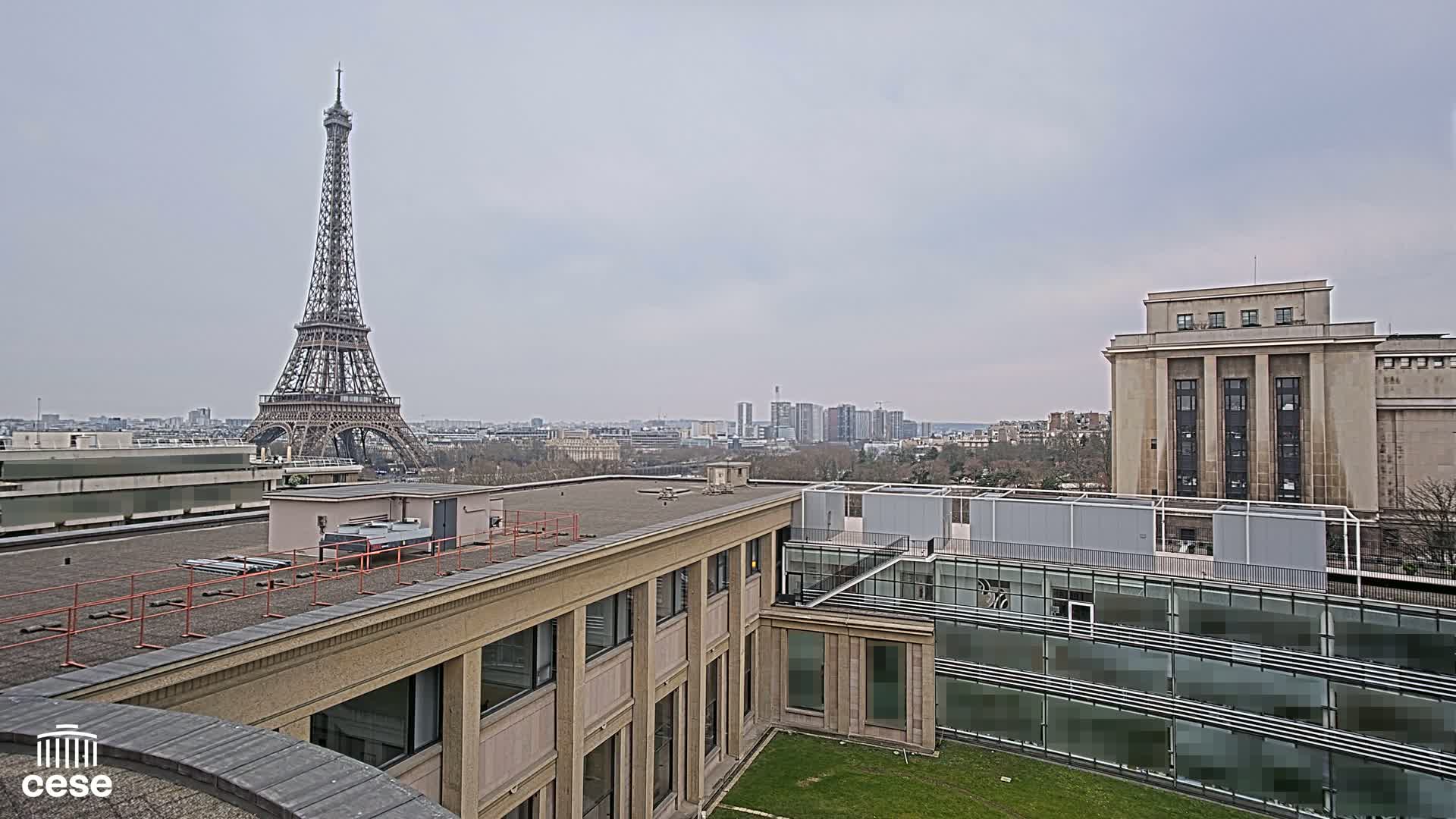 A view from a rooftop shows the Eiffel Tower in the distance under an overcast sky, with other buildings and a cityscape in the middle ground.