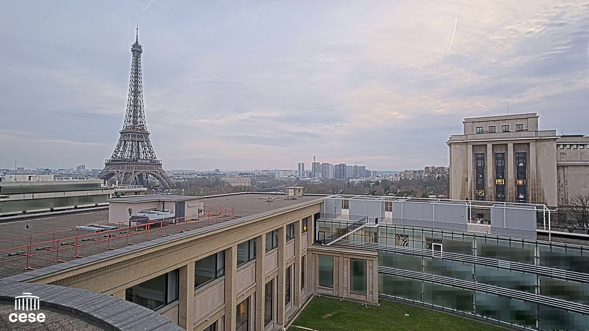 A rooftop view shows the Eiffel Tower in the distance under a cloudy sky, with modern and older buildings in the foreground.