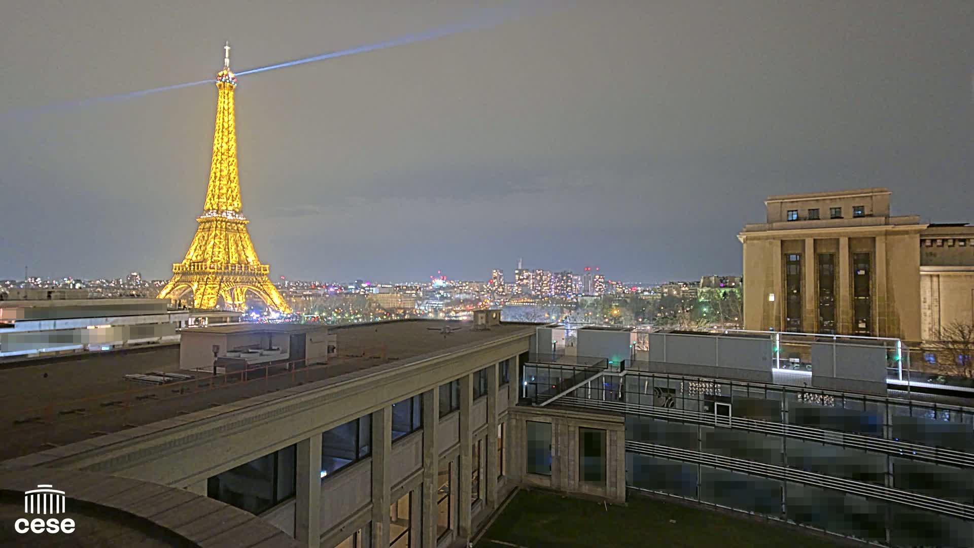 A nighttime view of the Eiffel Tower illuminated, with a city skyline and a large building in the foreground under a cloudy sky.