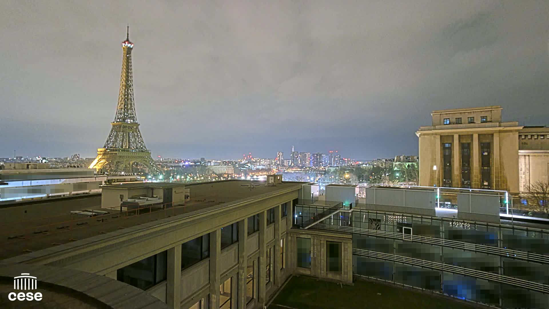 A nighttime view of the Eiffel Tower and a cityscape, seen from a rooftop overlooking another building under a cloudy sky.