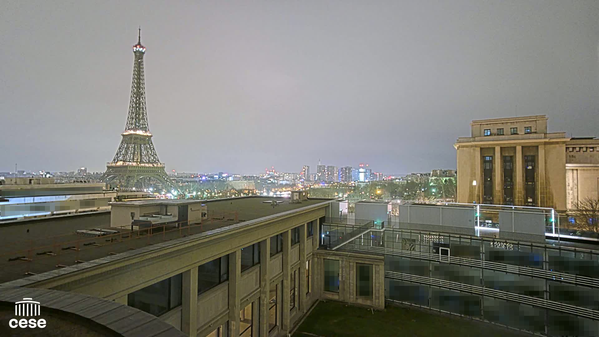 A nighttime view of the Eiffel Tower from a rooftop, with a city skyline and overcast skies in the background.