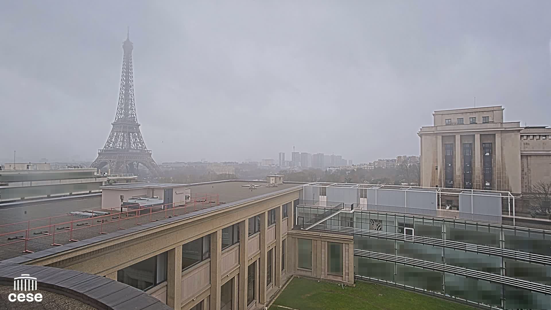 A foggy view of the Eiffel Tower from a rooftop, showing a cityscape and parts of beige buildings.