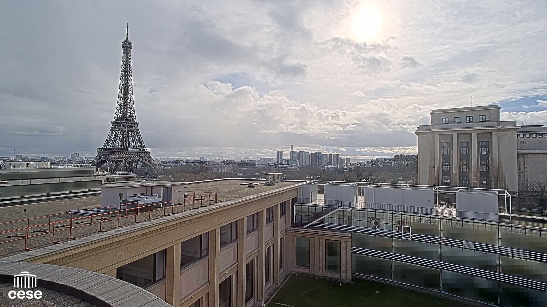 A partly sunny day shows the Eiffel Tower in the distance, viewed from a rooftop overlooking a city with several buildings, including a large, light-colored structure in the foreground.