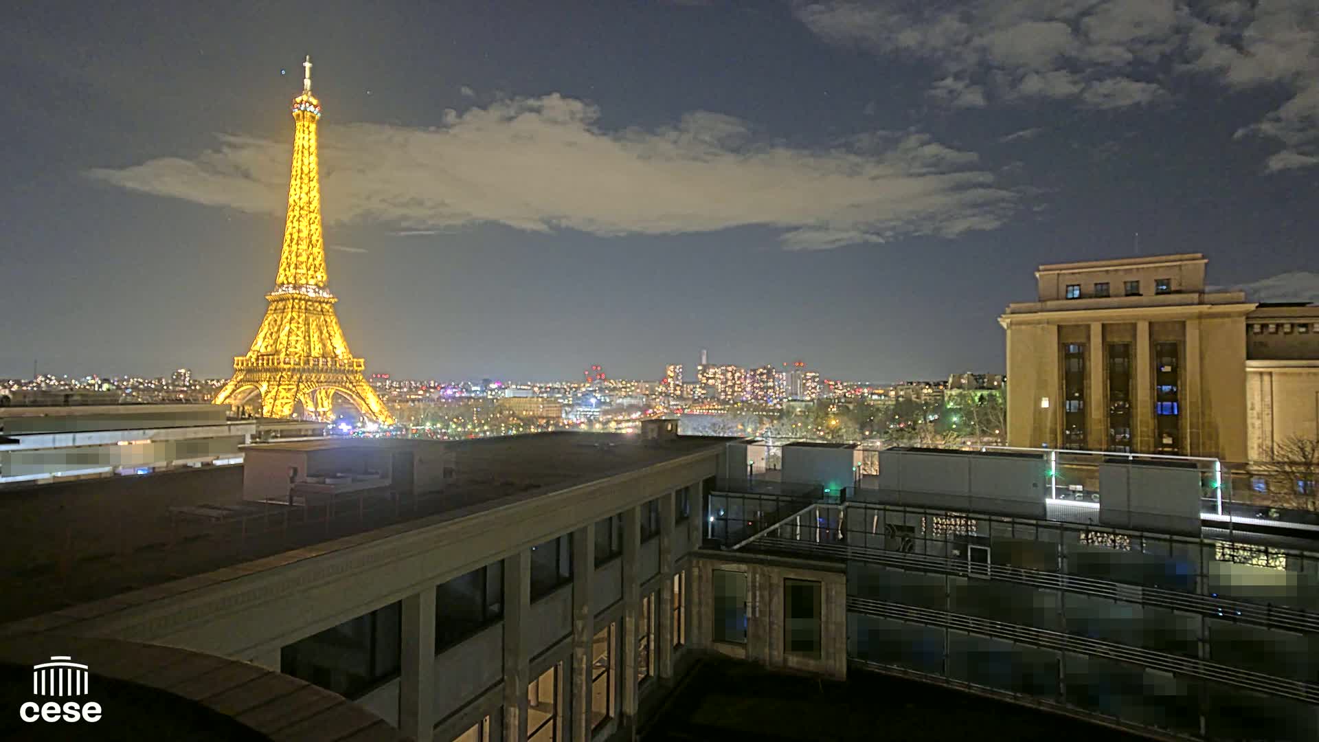 A nighttime view of the Eiffel Tower, illuminated in gold, overlooking a city skyline under a partly cloudy sky.