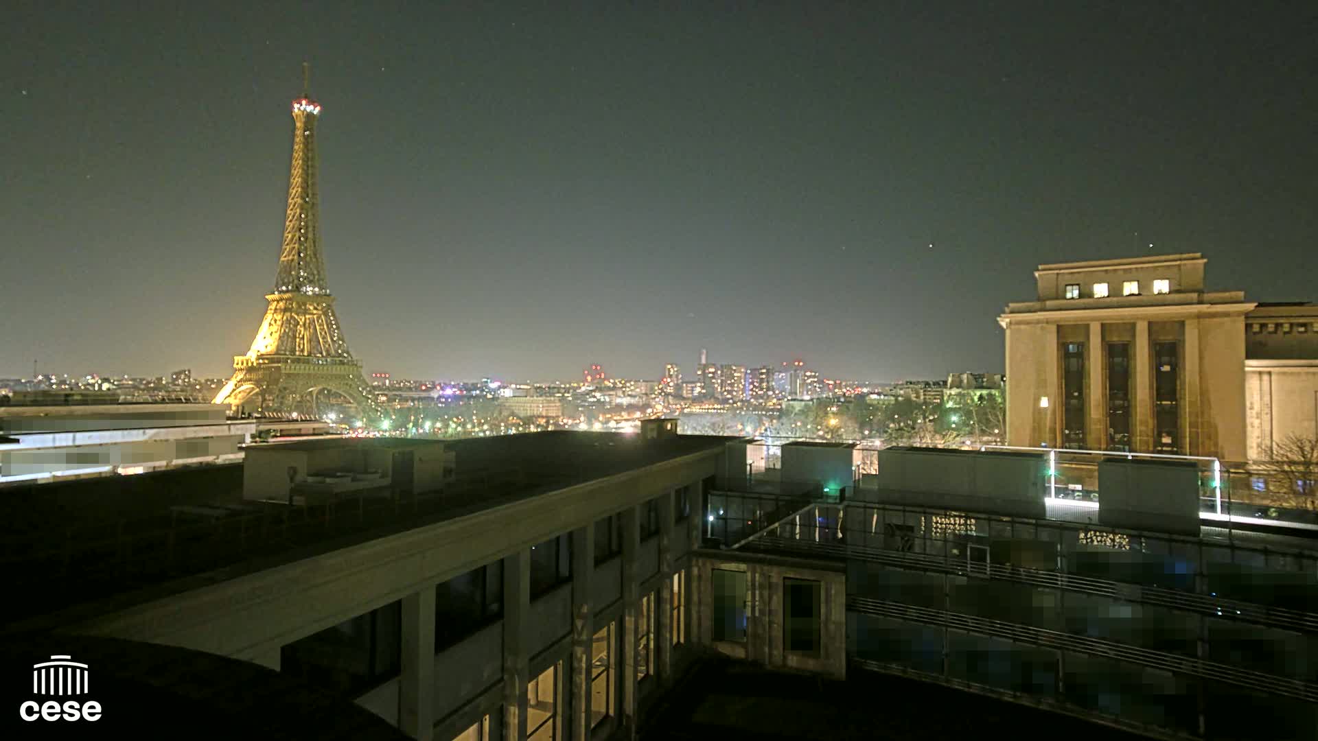 A nighttime view from a rooftop shows the Eiffel Tower illuminated against a dark sky, with a cityscape and a large building in the foreground.