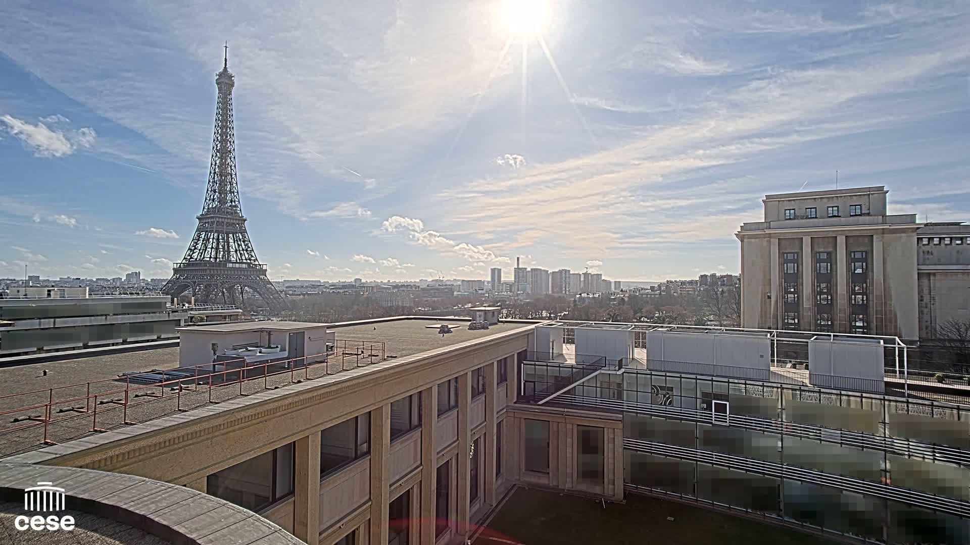 A sunny day view of the Eiffel Tower from a rooftop, overlooking other buildings and a cityscape.