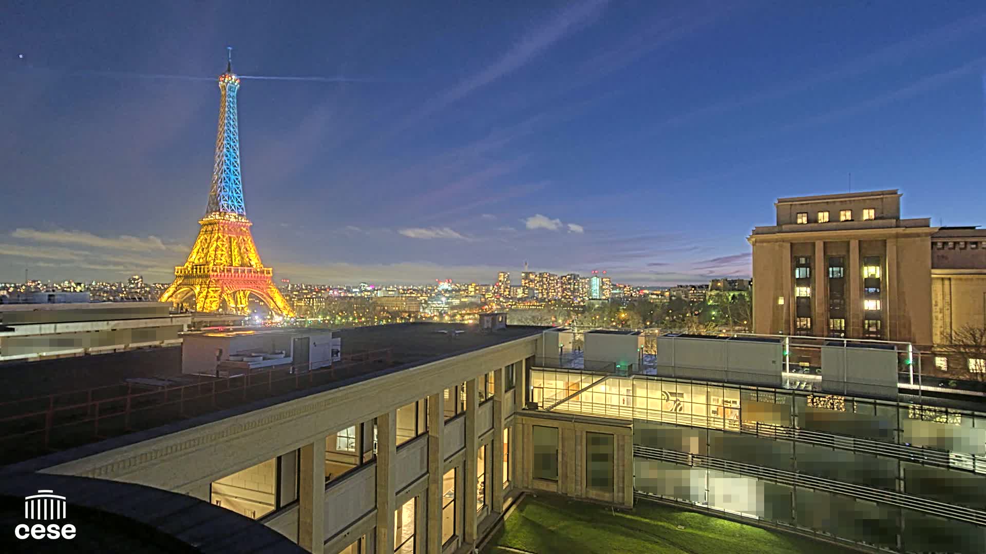 A brightly lit Eiffel Tower is visible at dusk from a rooftop overlooking a city skyline and a modern building.