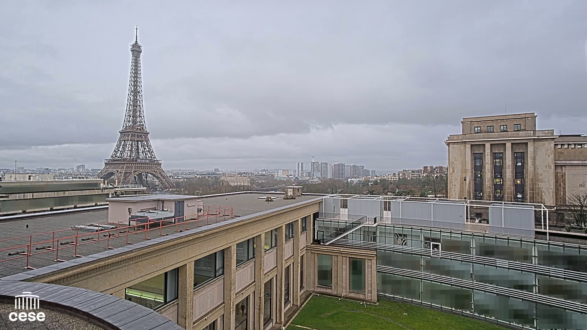 A rooftop view shows the Eiffel Tower in the distance under a cloudy sky, with modern and classical buildings in the foreground.