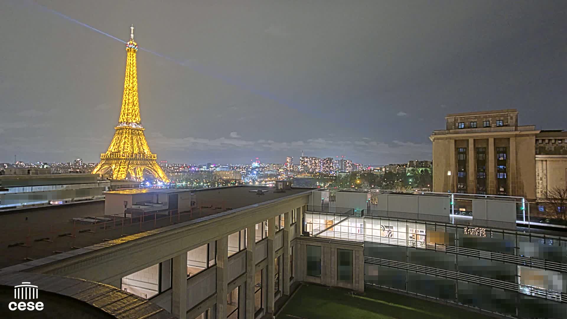 A nighttime view of the Eiffel Tower, illuminated in gold, from a rooftop overlooking a city skyline under a cloudy sky.