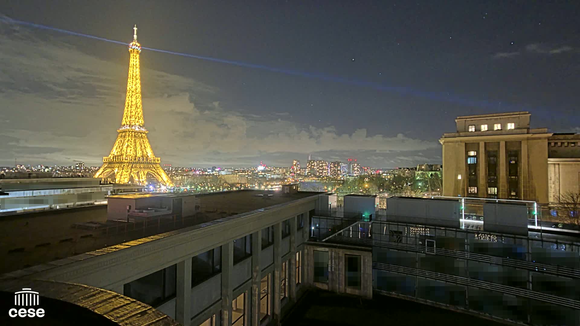 A nighttime view of the Eiffel Tower illuminated, with a cityscape and a building in the foreground under a partly cloudy sky.